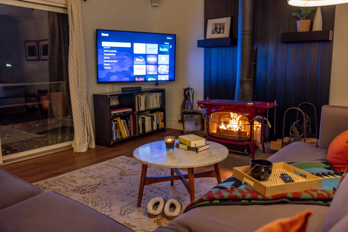 Evening view of the living room with a warm glow from the red wood-burning stove, a large flat-screen TV displaying streaming services, and a cozy purple sectional sofa.
