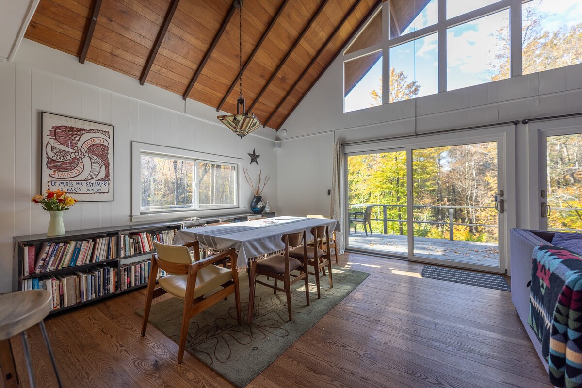 Dining area with a long wooden table and mid-century modern chairs, flanked by a low bookshelf, featuring large windows and sliding glass doors leading to a deck, all under a vaulted wood-paneled ceiling.