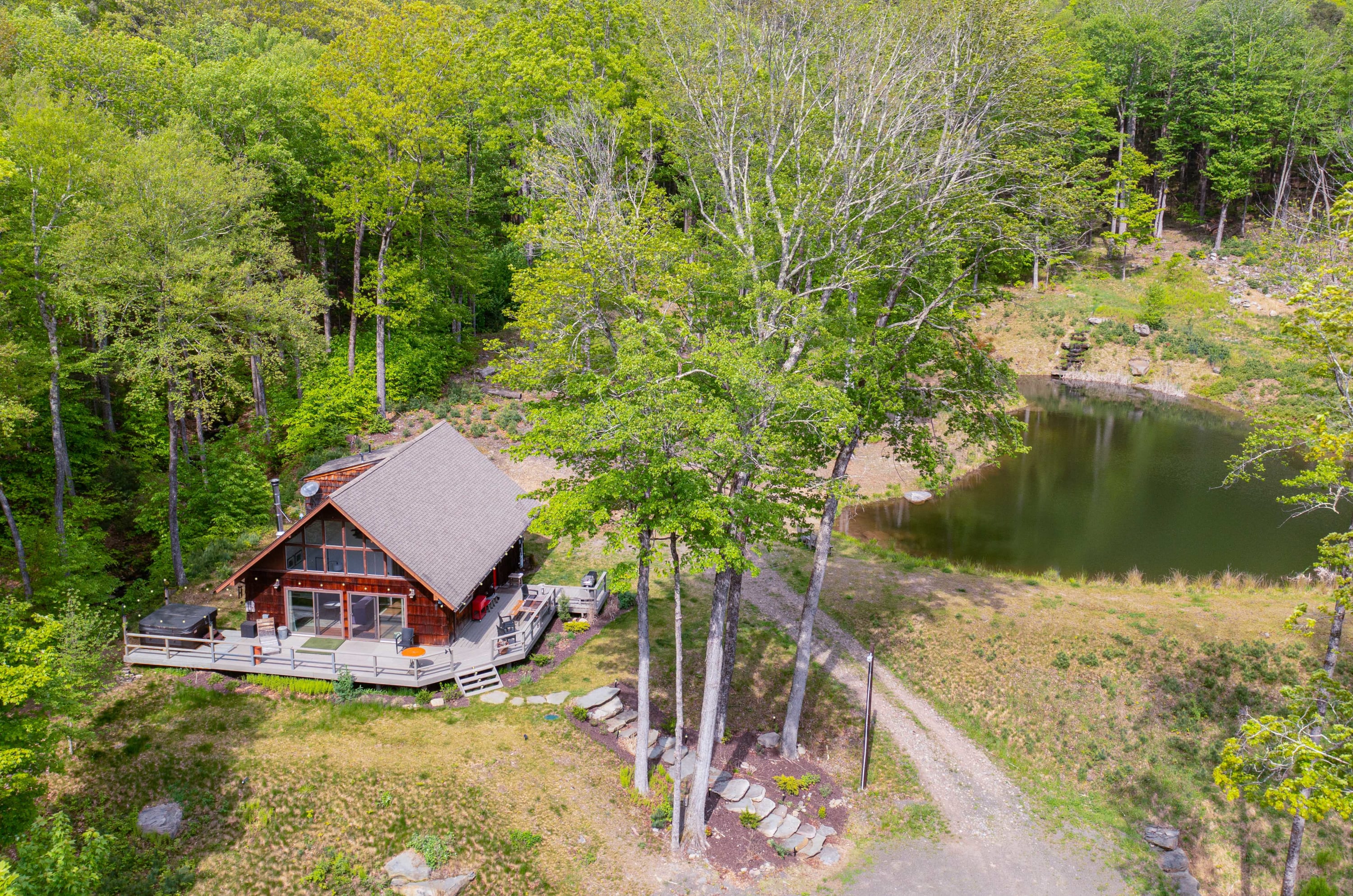An aerial shot reveals a modern A-frame style cabin with a large deck, situated next to a dark pond, with a dirt driveway leading up to it, all surrounded by vibrant green deciduous trees.