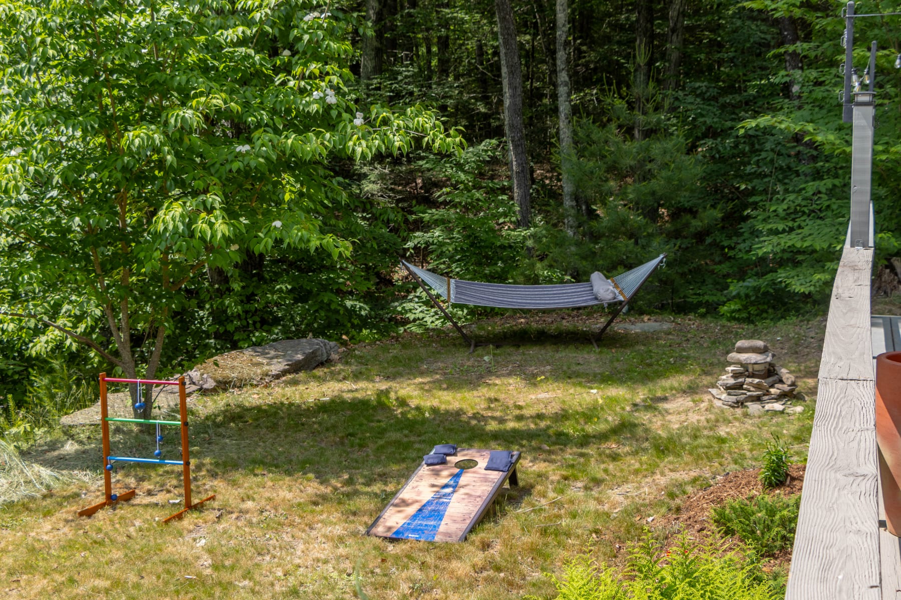 Grassy backyard area featuring a ladder toss game, a cornhole board, and a striped hammock strung between trees. A stone fire pit is also visible in the corner.