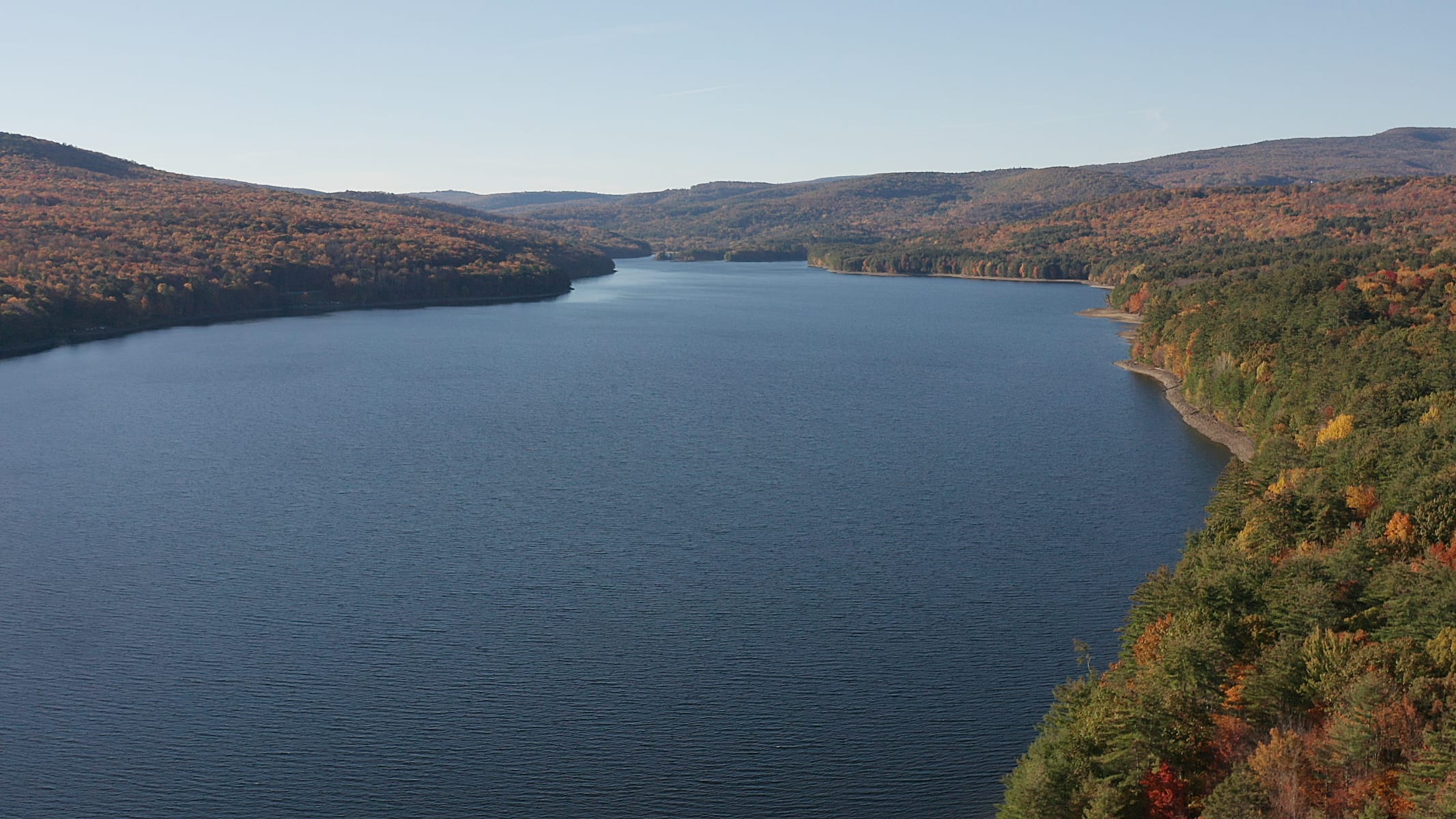 An expansive aerial view reveals a vast, deep blue lake winding through a landscape of rolling hills covered in dense forest, with patches of vibrant autumn foliage.