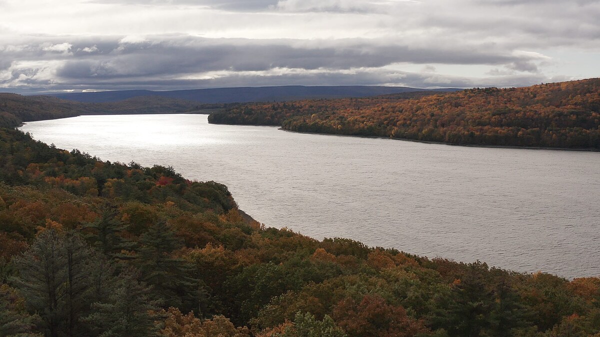 A wide, scenic view of a large lake with a bright reflection of the sky on its surface, bordered by hills covered in a mosaic of autumn-colored trees under a dramatic cloudy sky.