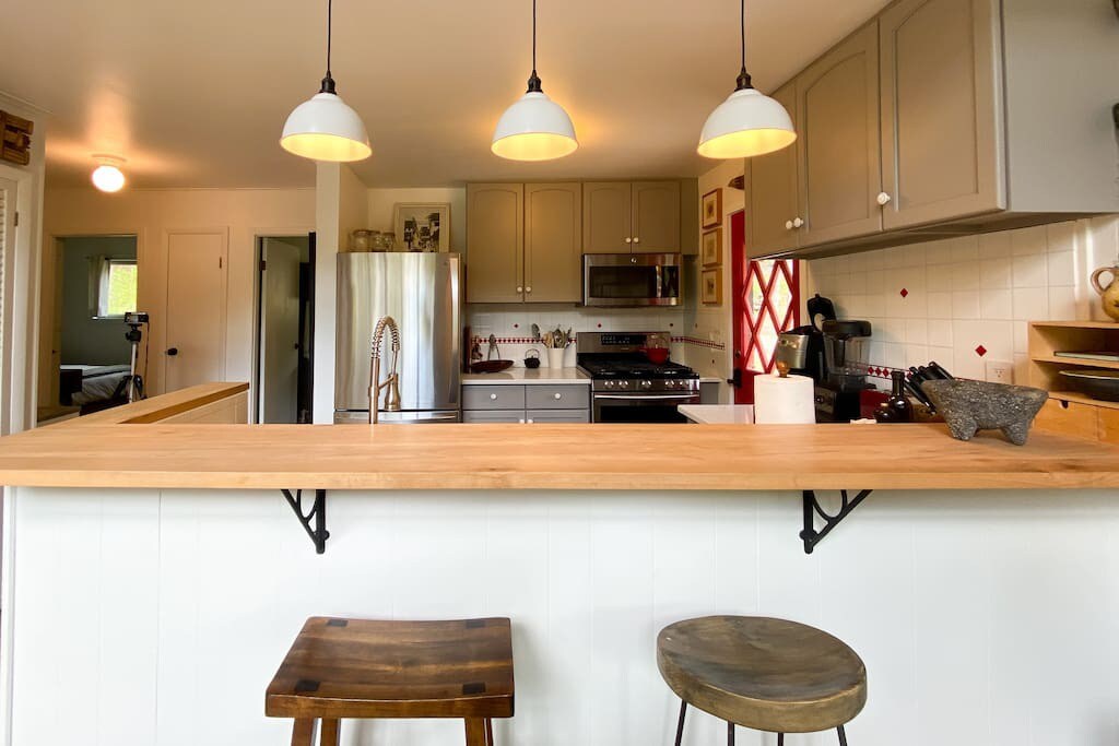Kitchen view highlighting a light wooden breakfast bar with two rustic bar stools, illuminated by three white pendant lights, with light gray cabinets and stainless steel appliances in the background.