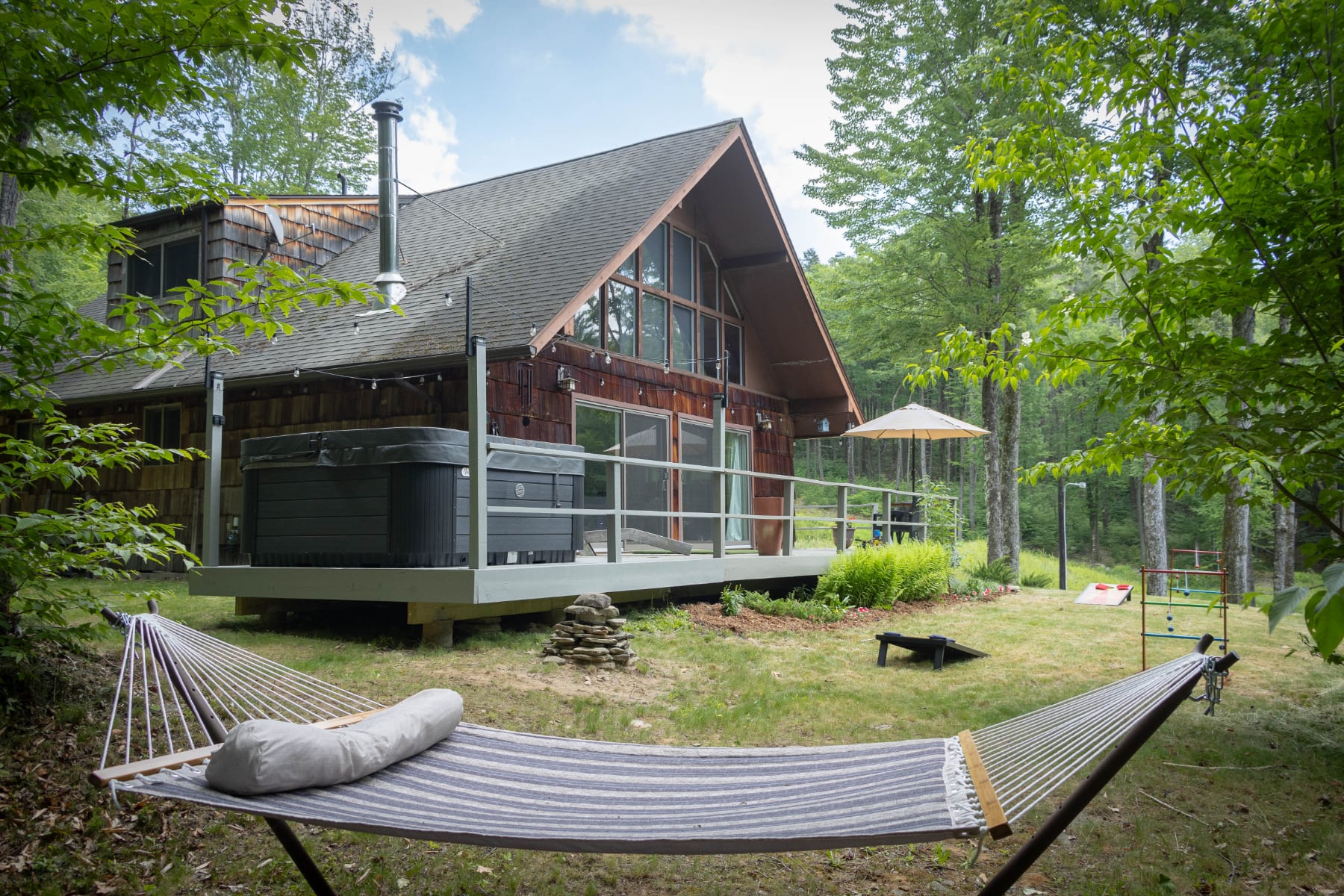 Rustic chalet-style house with a wooden exterior and large windows, surrounded by lush green trees. A spacious deck with a hot tub and outdoor seating overlooks a grassy yard with a hammock and lawn games.