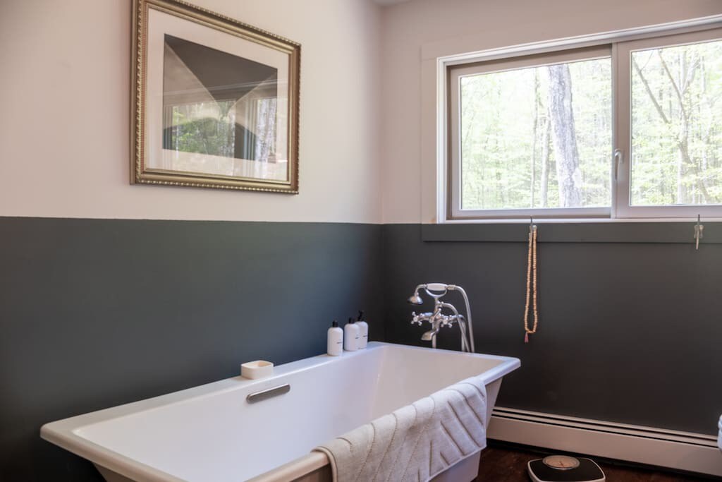 Close-up of the white freestanding bathtub in a bathroom with dark gray wainscoting, positioned beneath a large window offering a serene view of surrounding trees.