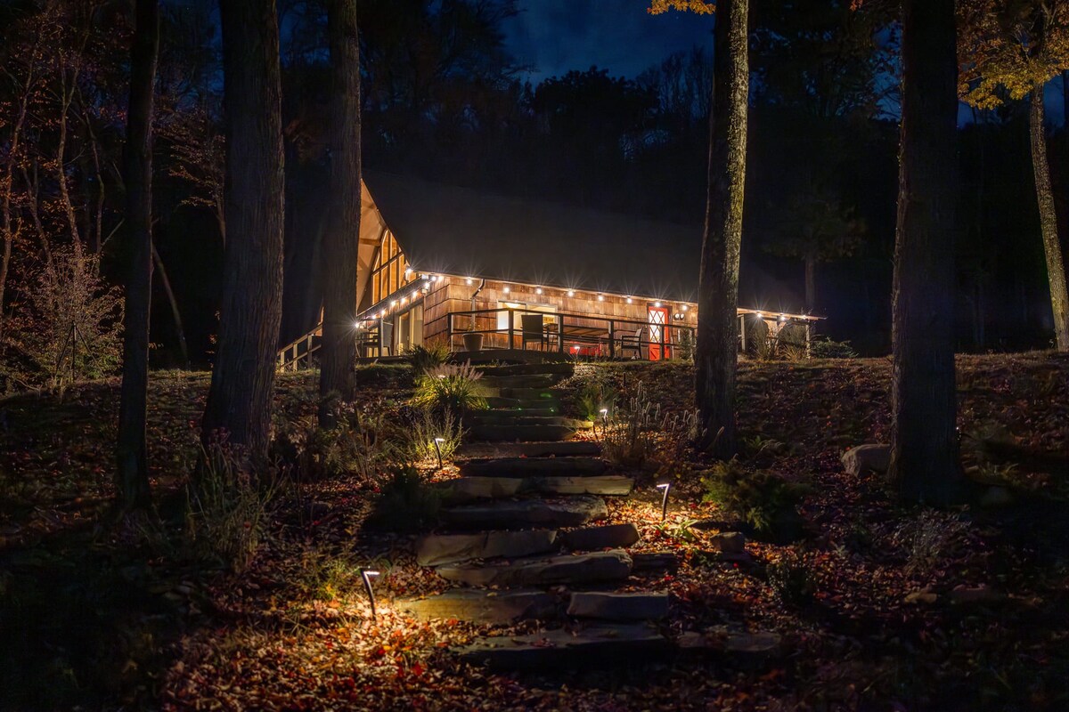 A rustic chalet-style house at night, illuminated by warm string lights along its wooden exterior and deck. A stone pathway with ground lights leads up to the house, surrounded by tall trees and fallen leaves, creating a cozy, inviting atmosphere.