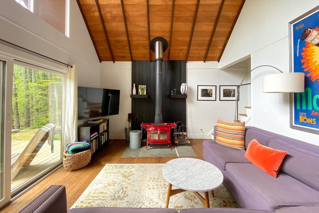 Cozy living room featuring a red wood-burning stove on a slate hearth, a purple sectional sofa, and a marble-top coffee table. Sliding glass doors open to the deck, and a TV is mounted on a bookshelf.