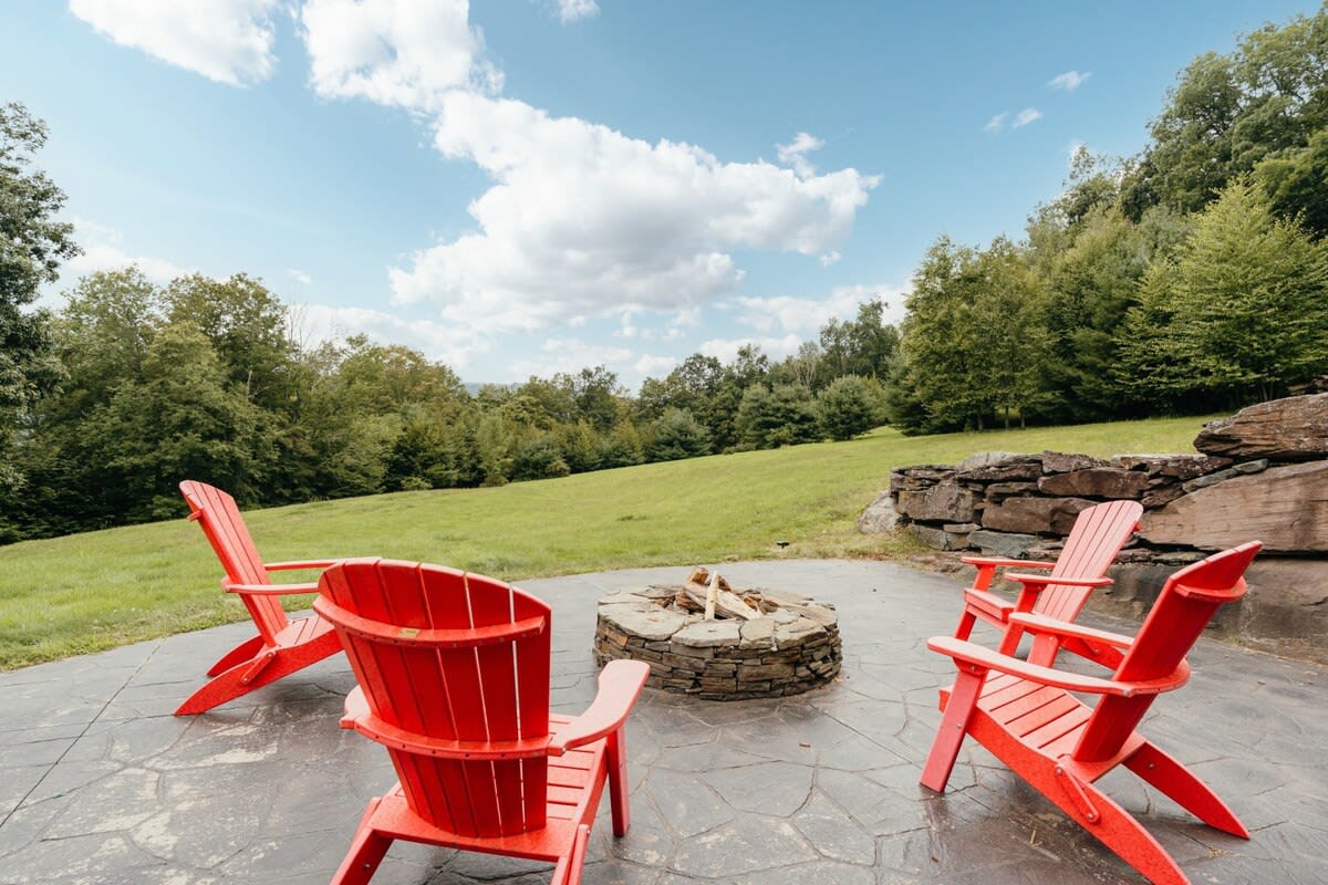 A stone fire pit with logs is surrounded by four bright red Adirondack chairs on a dark stamped concrete patio, overlooking a lush green lawn and forested hills under a blue sky.