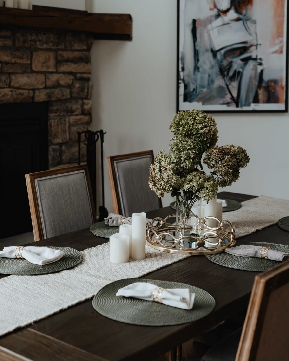 A close-up of a dark wooden dining table set with placemats, white napkins, and a centerpiece featuring candles and dried hydrangeas. A stone fireplace is visible in the background, along with a modern abstract painting on the wall.