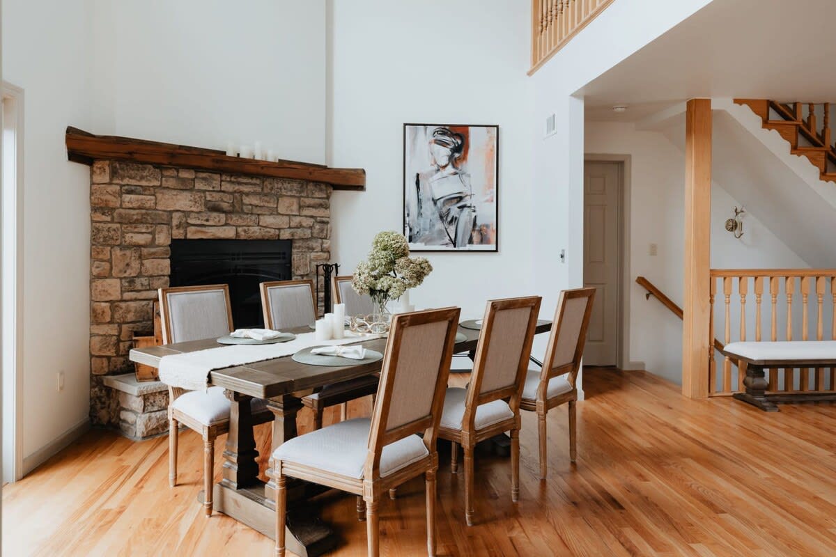 A bright dining room with a large wooden table and eight upholstered chairs, set against a stone fireplace with a rustic wooden mantel. A modern abstract painting hangs on the white wall, and hardwood floors extend throughout the space.