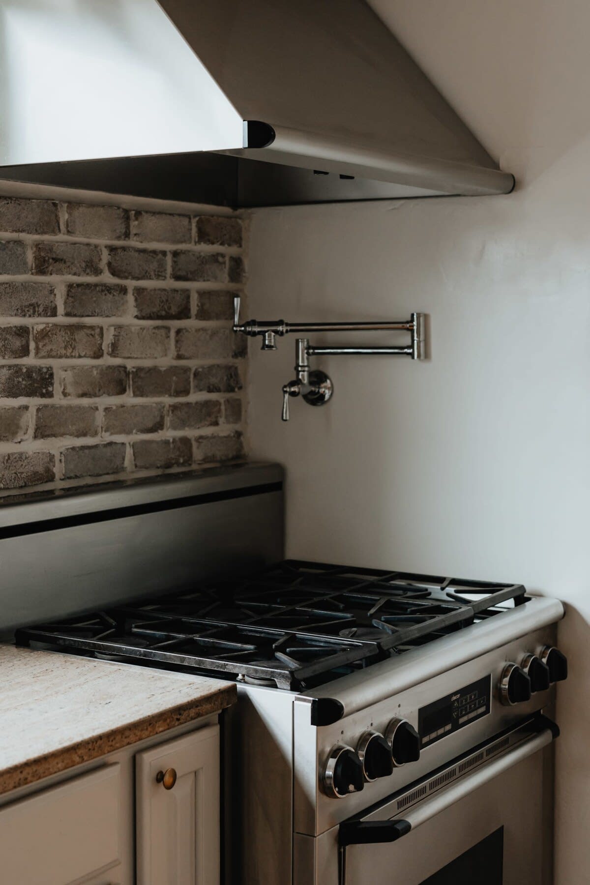 Close-up of a stainless steel gas range with black grates, a pot filler faucet on a brick backsplash, and a stainless steel range hood. A light-colored countertop is visible on the left.