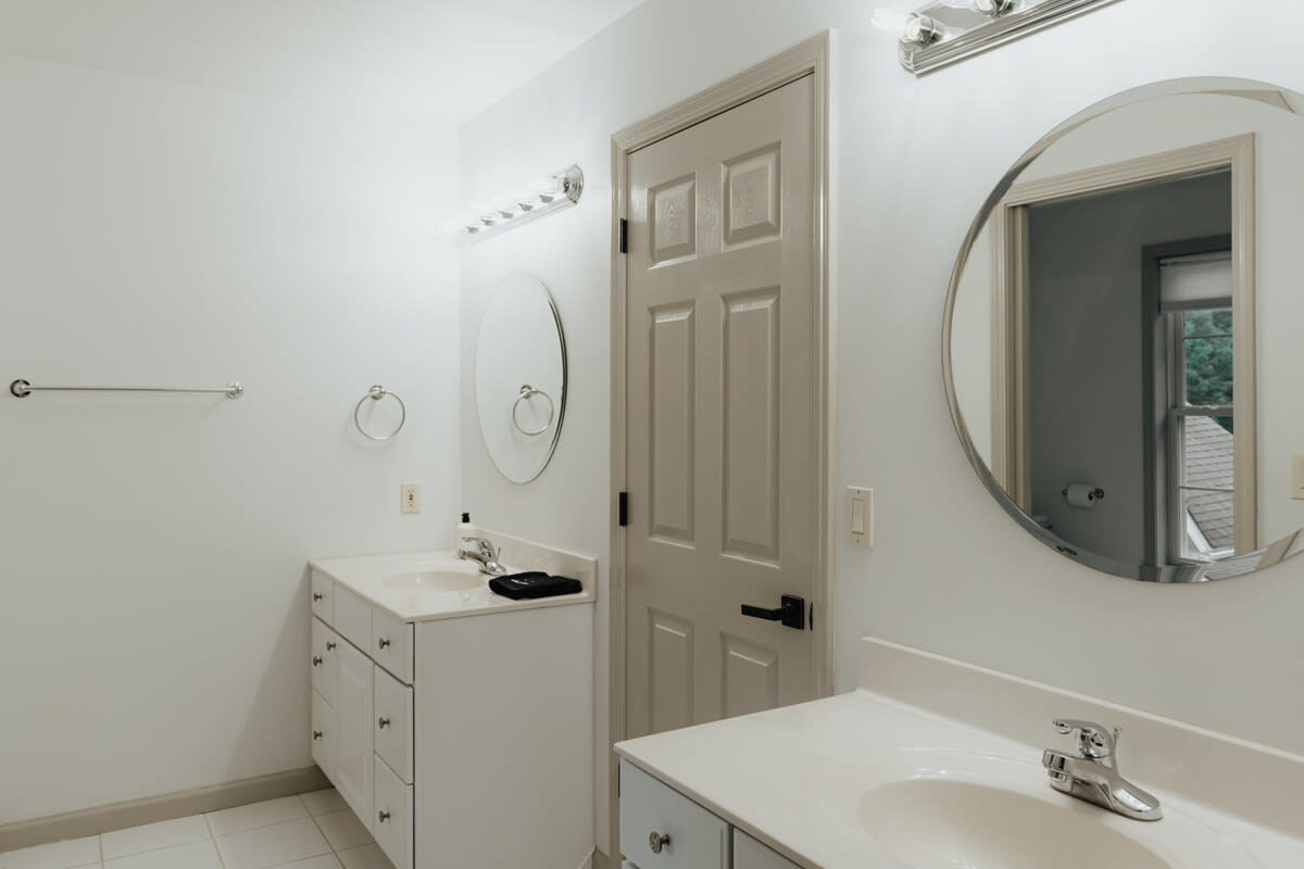 A spacious bathroom with two white vanity sinks, large round mirrors, and chrome light fixtures above each. A light-colored door is centered between the vanities, and the floor is tiled.