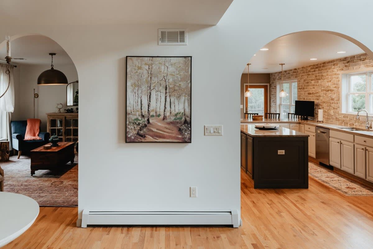 An interior view showcasing two arched doorways, one leading to a cozy living room with a dark sofa, and the other opening into a kitchen with a large island and brick backsplash, all on hardwood floors.