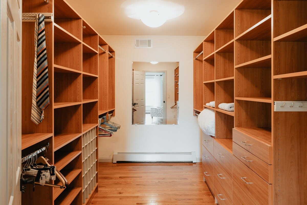 A large walk-in closet featuring extensive light wood built-in shelving units and drawers on both sides, with a full-length mirror at the far end reflecting an open doorway.
