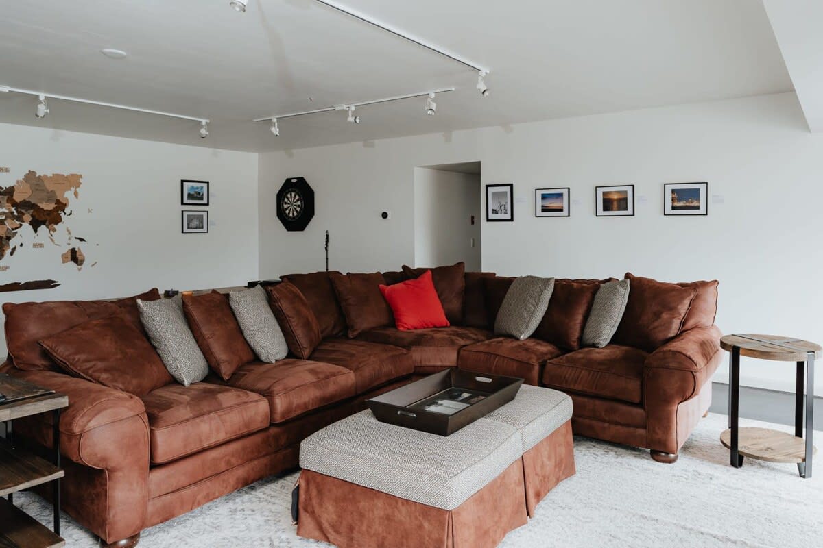 A wide shot of a large living room featuring a brown sectional sofa, an ottoman, and a white area rug, with a wooden world map and dartboard visible on the far wall.