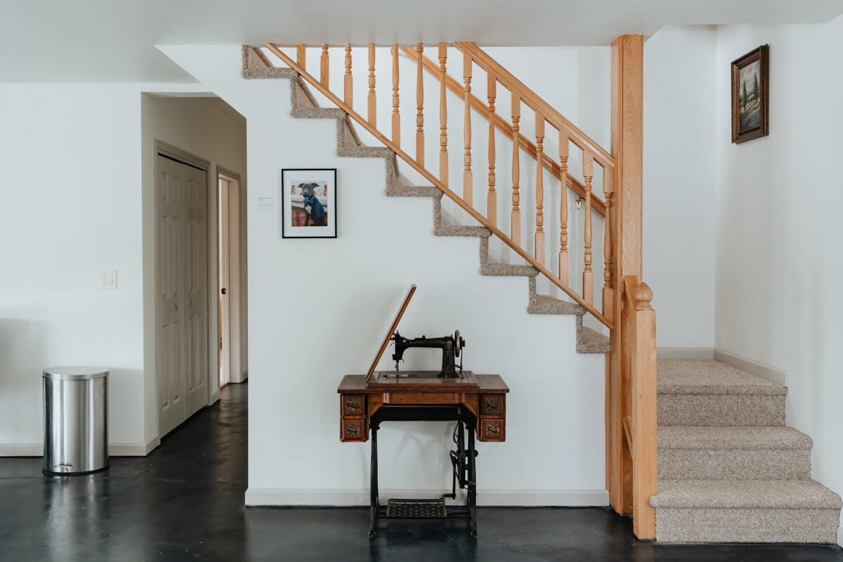 A welcoming foyer area featuring a carpeted staircase with a light wood banister, an antique sewing machine on a wooden table, and a framed dog portrait on the wall.