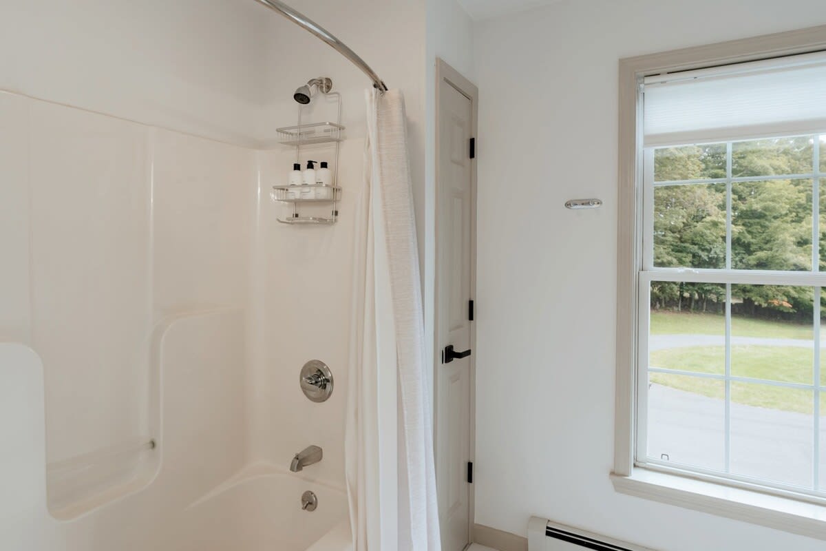 A clean bathroom featuring a white shower-tub combination with a chrome showerhead and a caddy holding toiletries. A window with a view of lush greenery is visible next to the shower.