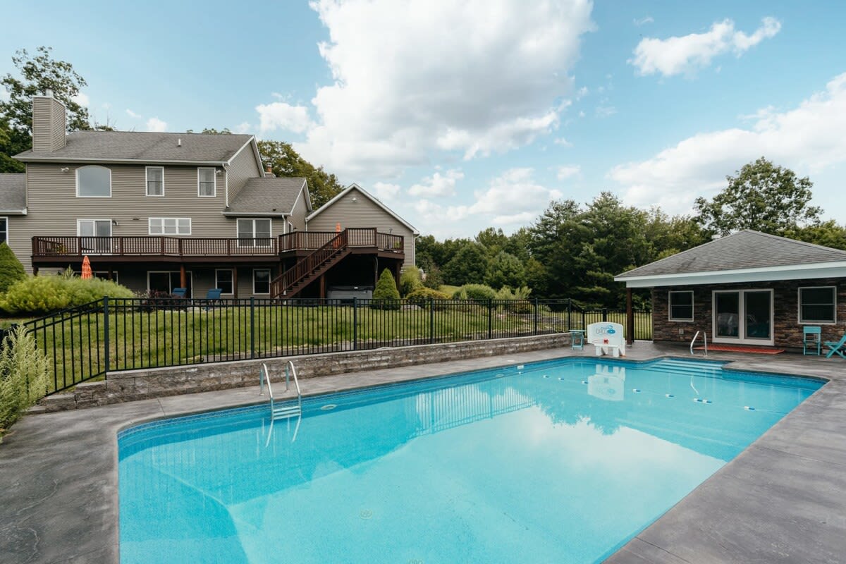 A large rectangular swimming pool with steps and a ladder is set on a concrete patio, with a two-story house featuring a multi-level deck and a separate pool house visible in the background, surrounded by green hills.