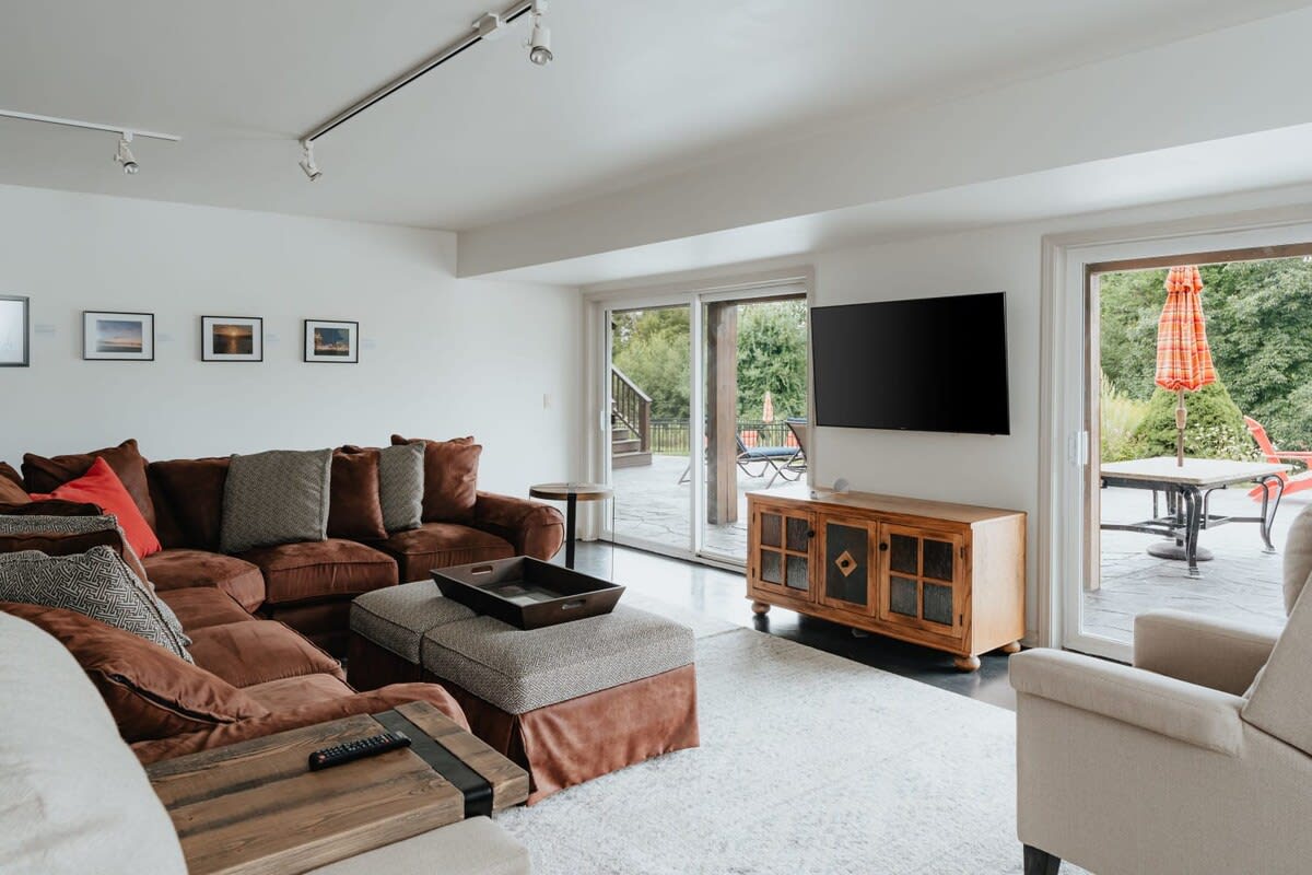 A cozy living room features a large sectional sofa in a warm brown tone, a flat-screen TV mounted on the wall, and sliding glass doors leading to an outdoor patio. A light rug covers the dark floor, and track lighting illuminates the space.