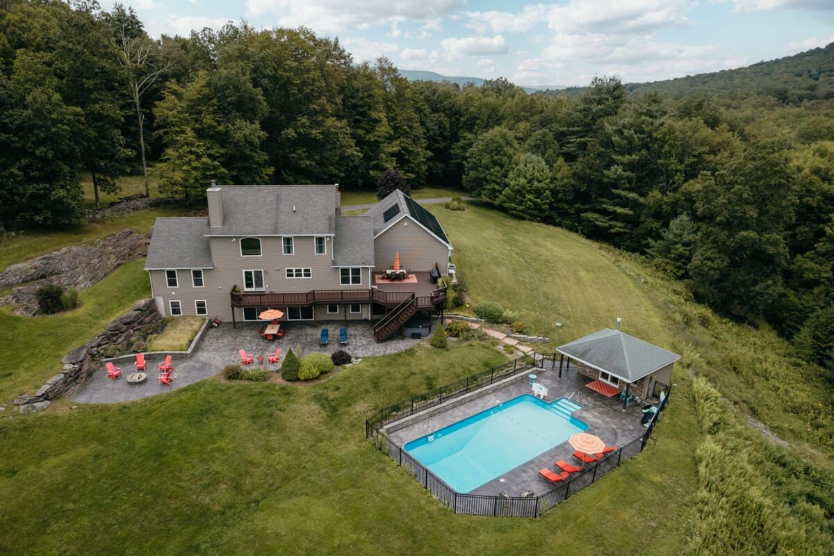 An aerial view showcases a large house with a multi-level deck, a fenced inground swimming pool, and a pool house, all set on a grassy hillside surrounded by dense green trees and distant mountains.