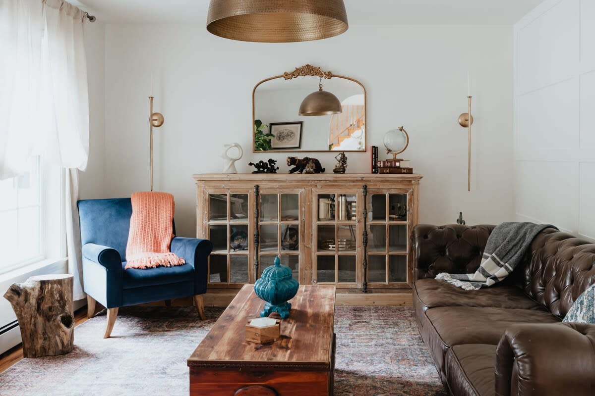 A cozy living room featuring a dark brown leather Chesterfield sofa, a blue velvet armchair with a peach throw, and a rustic wooden coffee table. A large wooden cabinet with glass doors and a gold mirror above it adorns the back wall, under a large brass pendant light.