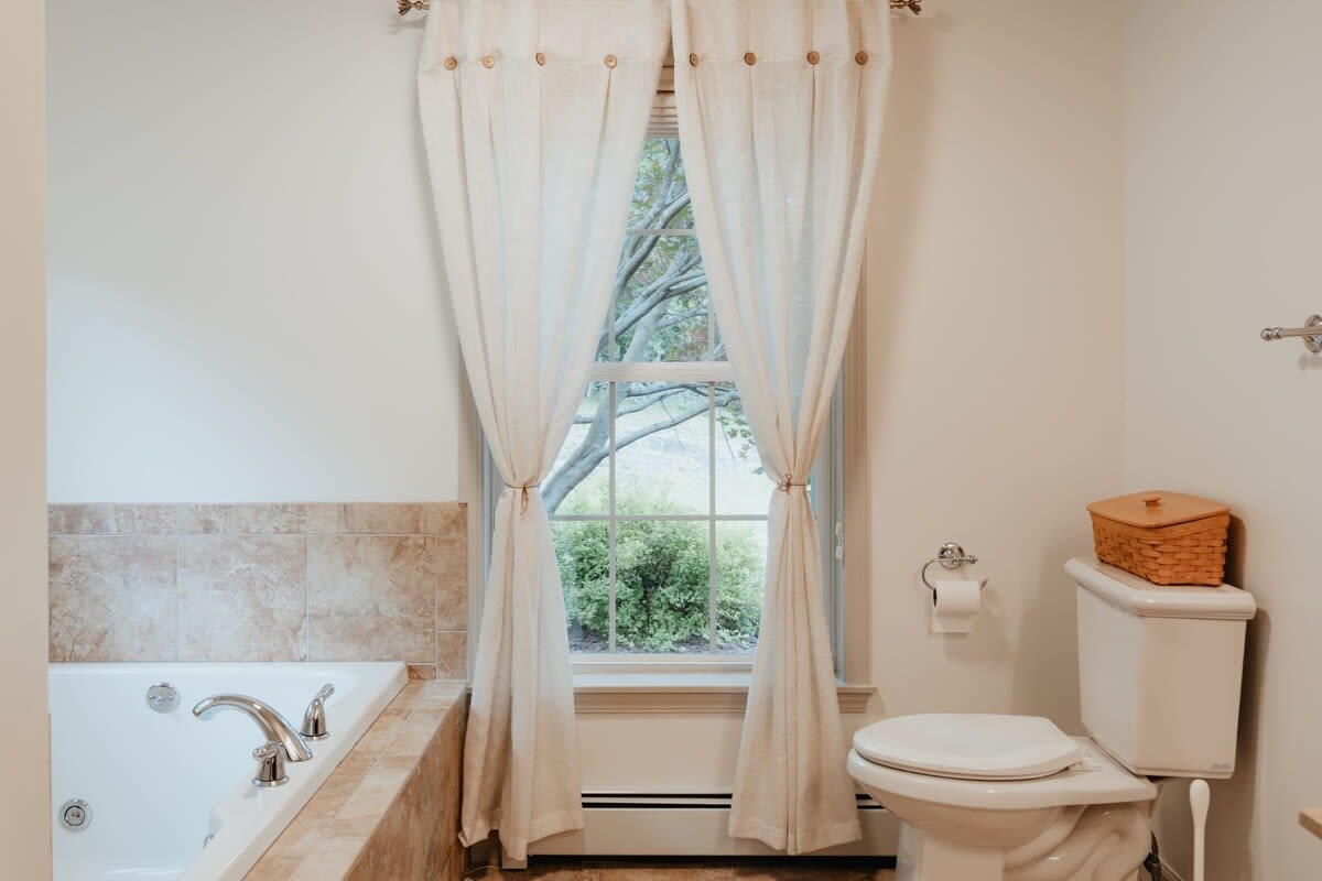 A bright bathroom featuring a jetted bathtub with light brown tiled surround, a toilet, and a large window with light-colored button-tab curtains tied back, offering a view of trees outside.