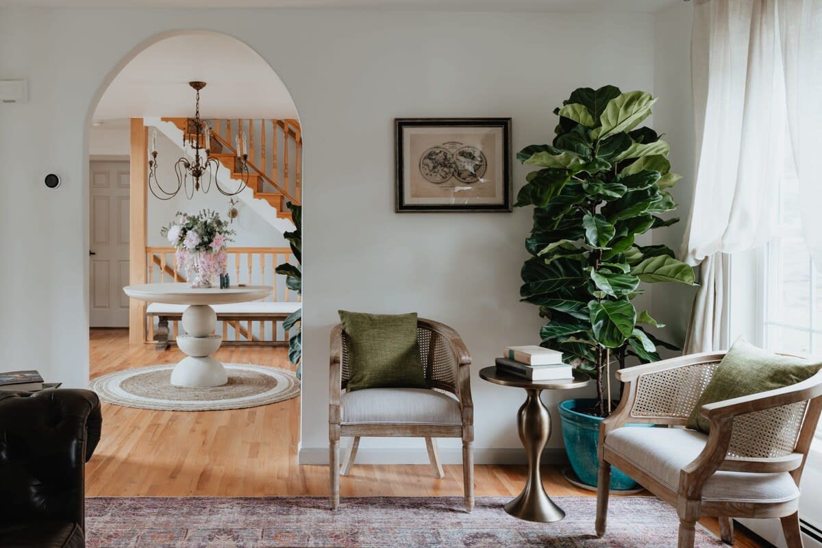 A cozy living space featuring two cane-back armchairs with green pillows, a fiddle leaf fig plant, and a patterned rug, with an arched doorway leading to a bright foyer with a round table and staircase.