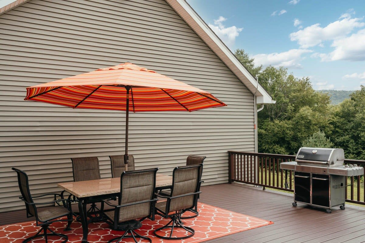 An outdoor deck with a brown slatted floor, featuring a dining table with four chairs under a large orange striped umbrella. A stainless steel BBQ grill is positioned nearby, ready for outdoor cooking.