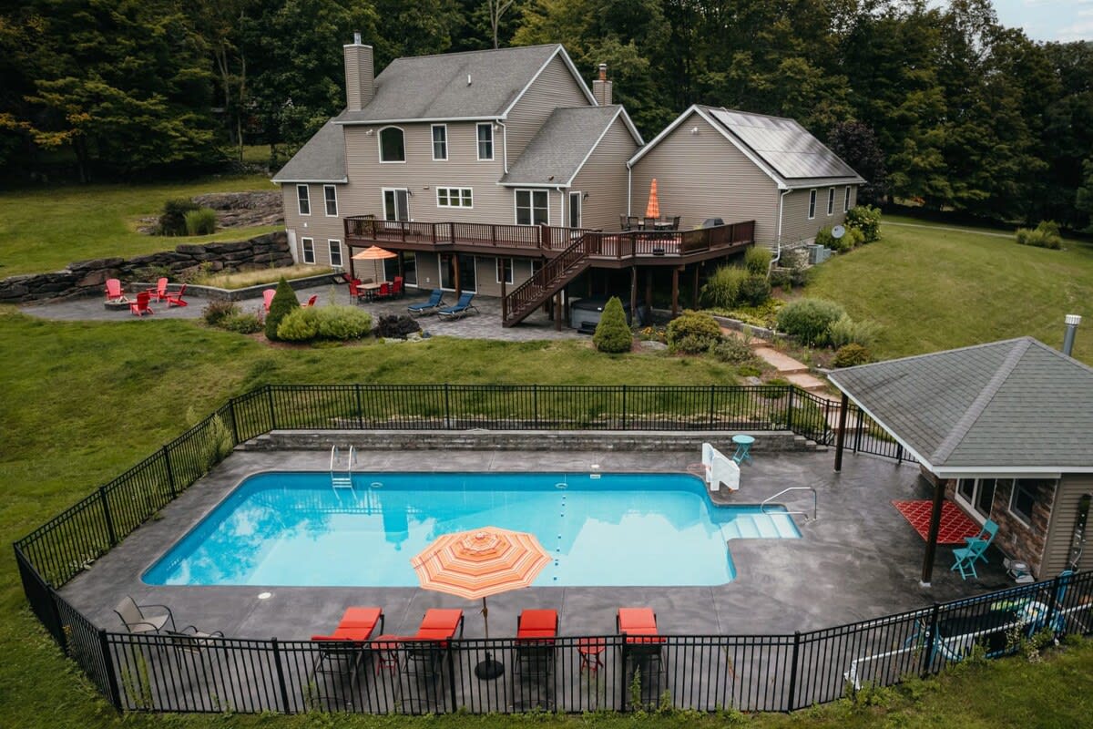 An aerial shot captures the large house with its multi-level deck, a fenced inground swimming pool, and a pool house, all nestled on a green hillside surrounded by lush trees and distant mountains.