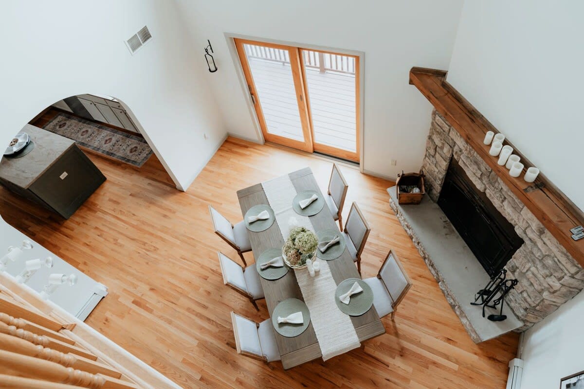 An aerial view of the dining room, showcasing the large wooden table with eight chairs, a stone fireplace, and hardwood floors. Large glass doors lead to an outdoor deck, and a glimpse of the kitchen is visible through an archway.