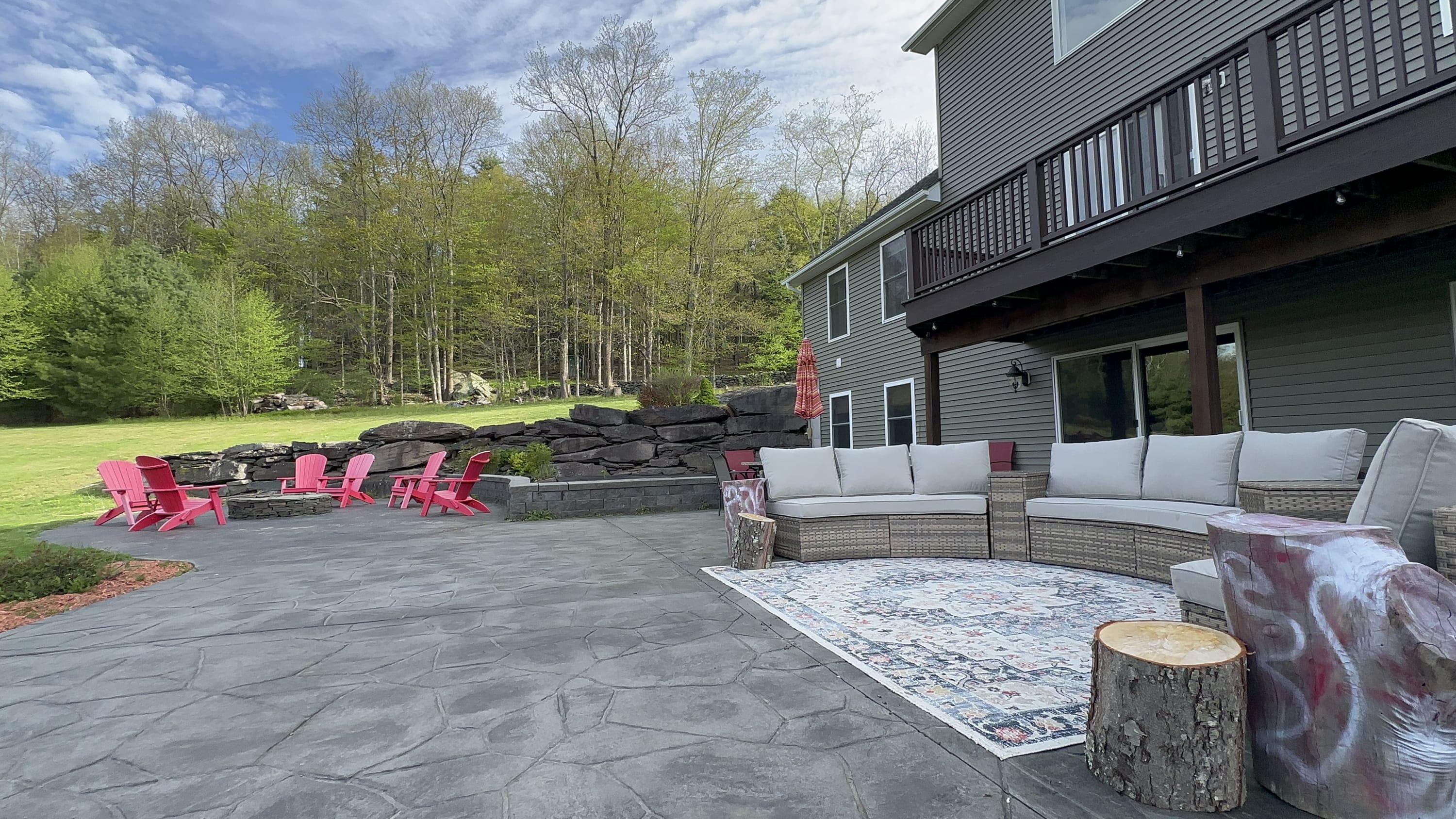 A large stamped concrete patio with a curved wicker sectional sofa and a decorative outdoor rug. A stone fire pit area with red Adirondack chairs is visible in the background, surrounded by a grassy hill.