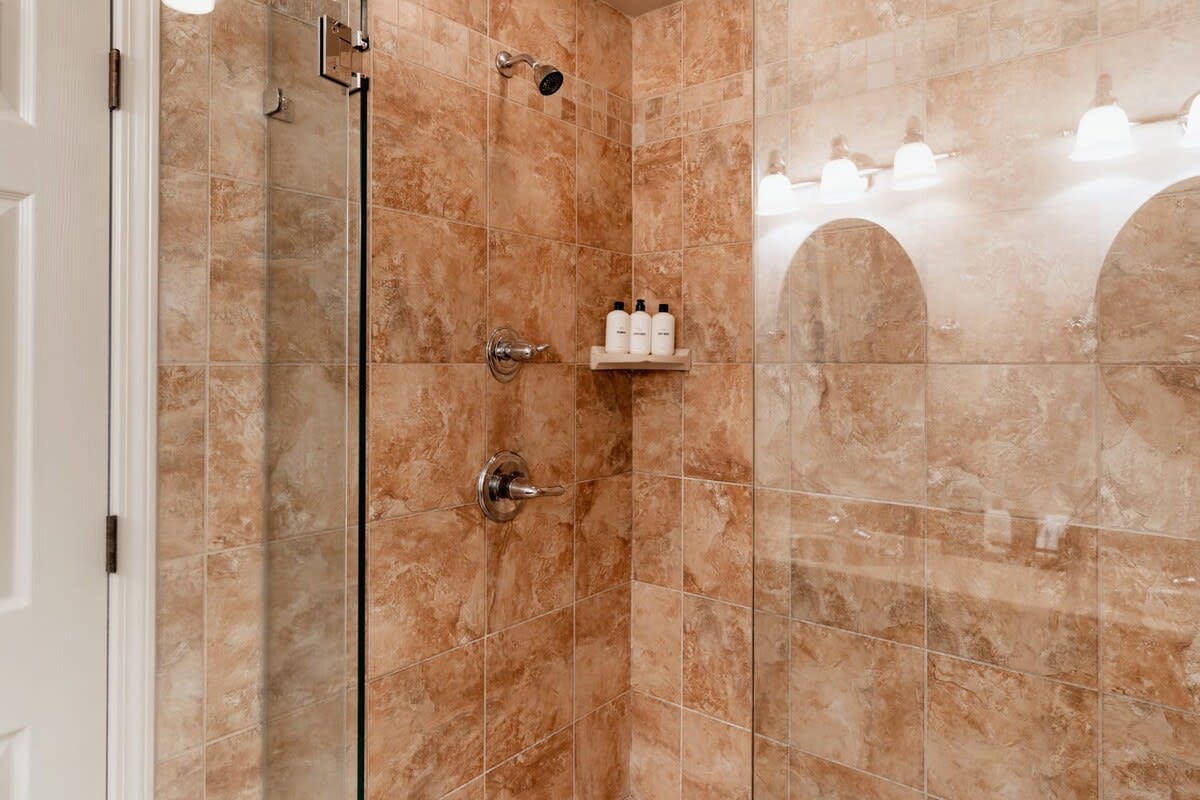 A spacious walk-in shower with warm brown tiled walls and floor, featuring a clear glass enclosure, a rain shower head, and a small wooden shelf with toiletries.