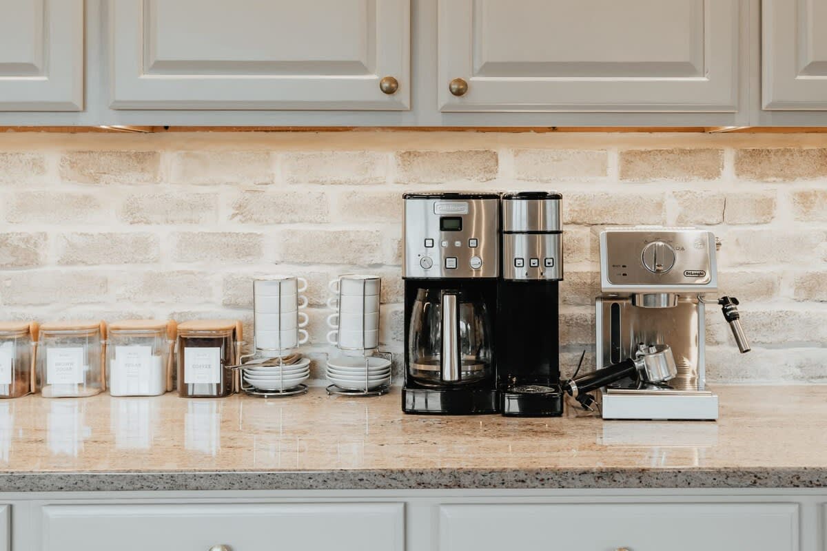 A close-up of a well-equipped coffee station in a kitchen, featuring a drip coffee maker, an espresso machine, and neatly organized cups and canisters, set against a light brick backsplash and granite countertop.