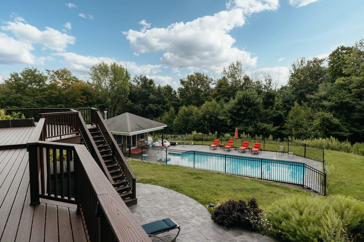 An expansive outdoor scene showcasing a fenced swimming pool with lounge chairs, a covered cabana, and a multi-level deck leading down from the house. Lush green trees surround the property.