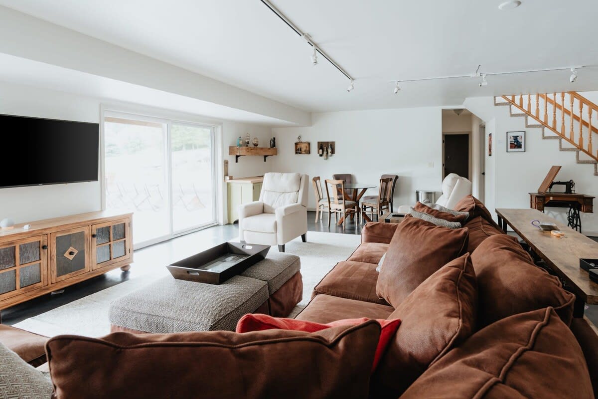 A spacious living room with a large brown sectional sofa, a TV on a wooden console, and a dining area with a glass table, featuring a sliding glass door leading to an outdoor patio.