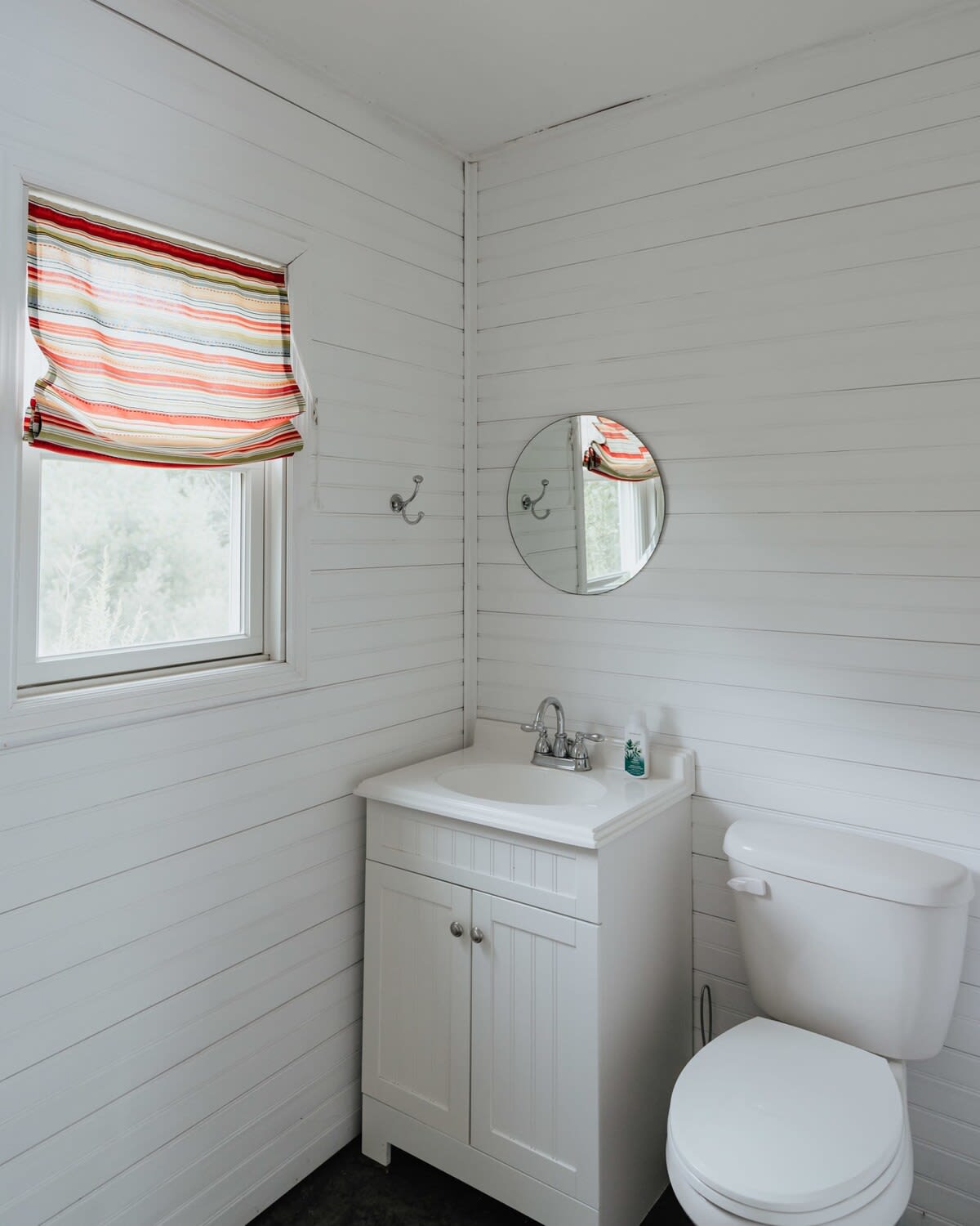 A compact bathroom with white shiplap walls, a white vanity with a sink, a toilet, and a small round mirror. A window with a colorful striped Roman shade adds a pop of color.