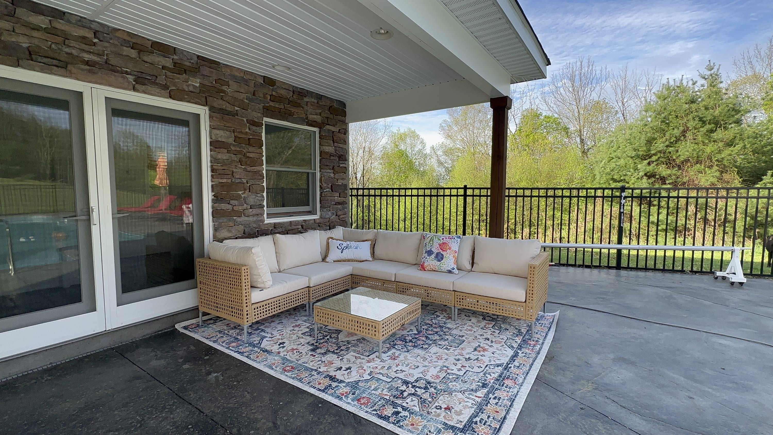 A covered patio features a light-colored wicker sectional sofa with cream cushions and a glass-top coffee table, all placed on a patterned rug, with a stone wall and large glass doors leading indoors.