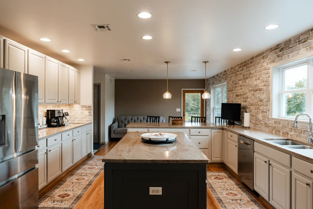 Wide shot of a modern kitchen featuring light gray shaker cabinets, stainless steel appliances, a large island with a dark base and light granite countertop, and a brick accent wall with windows. Two pendant lights hang over the island.