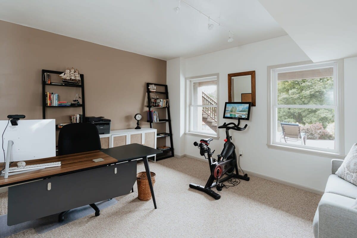 A bright office space with a large desk, black office chair, and shelving units, also featuring a Peloton bike, all set on light carpet with large windows overlooking greenery.