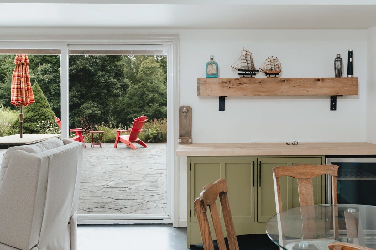 A wet bar area with green cabinets, a wooden countertop, and a rustic floating shelf adorned with decorative boats, offering a view through a sliding door to an outdoor patio with red Adirondack chairs.