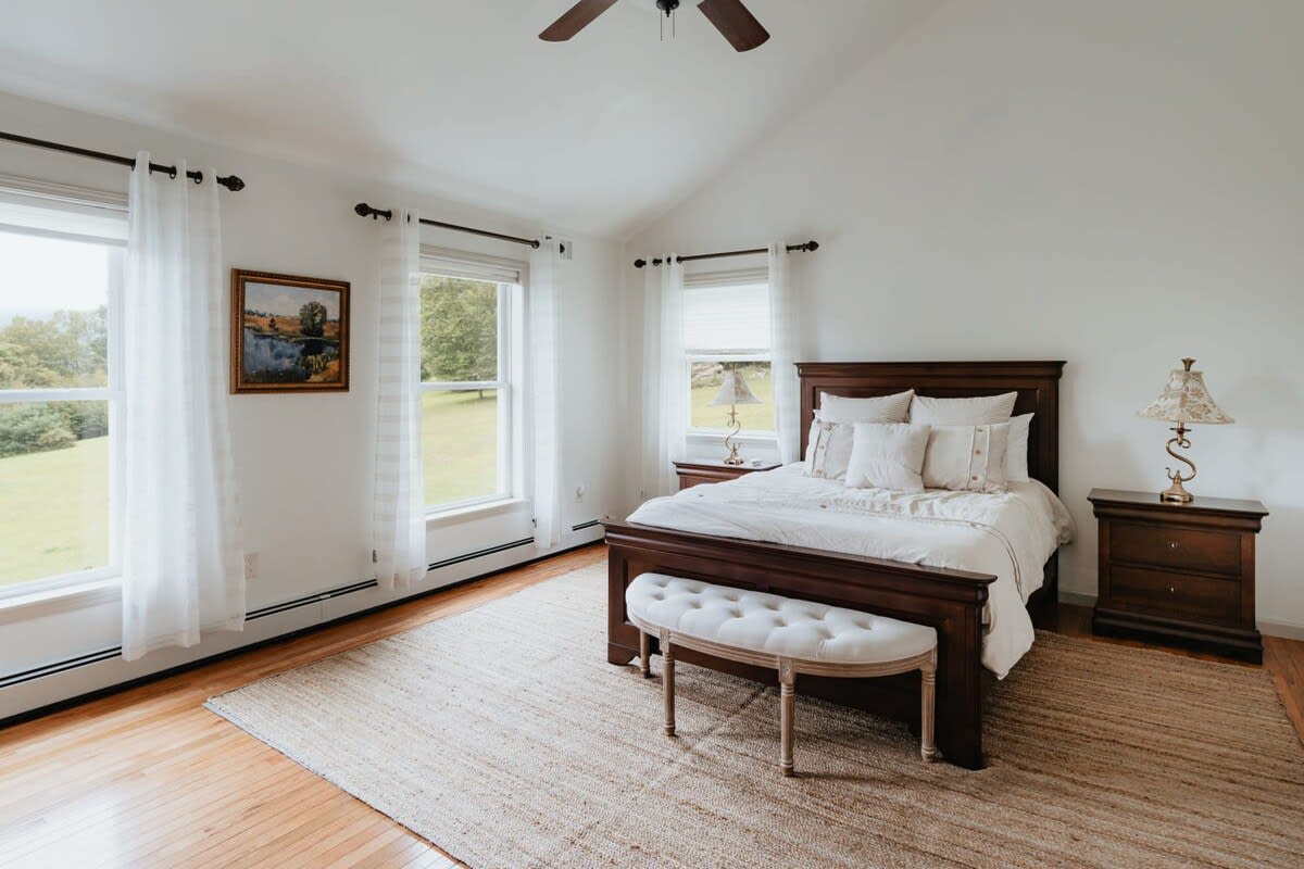 A bright bedroom with a dark wooden bed frame, a white upholstered bench at the foot, and two matching nightstands with lamps. Large windows offer views of the outdoors, and a jute rug covers the hardwood floor.