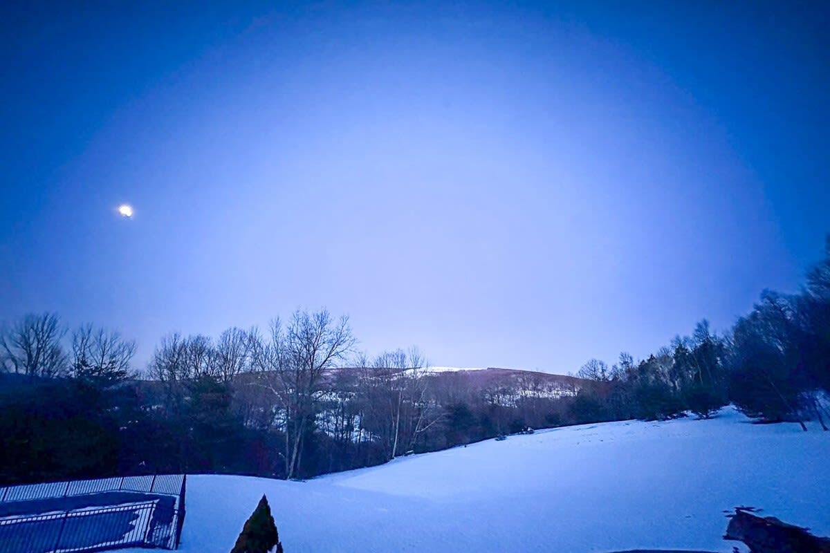 A wide-angle view of a snow-covered landscape under a dark blue twilight sky, with a bright moon, showing distant hills and trees, and a fenced pool area in the foreground.