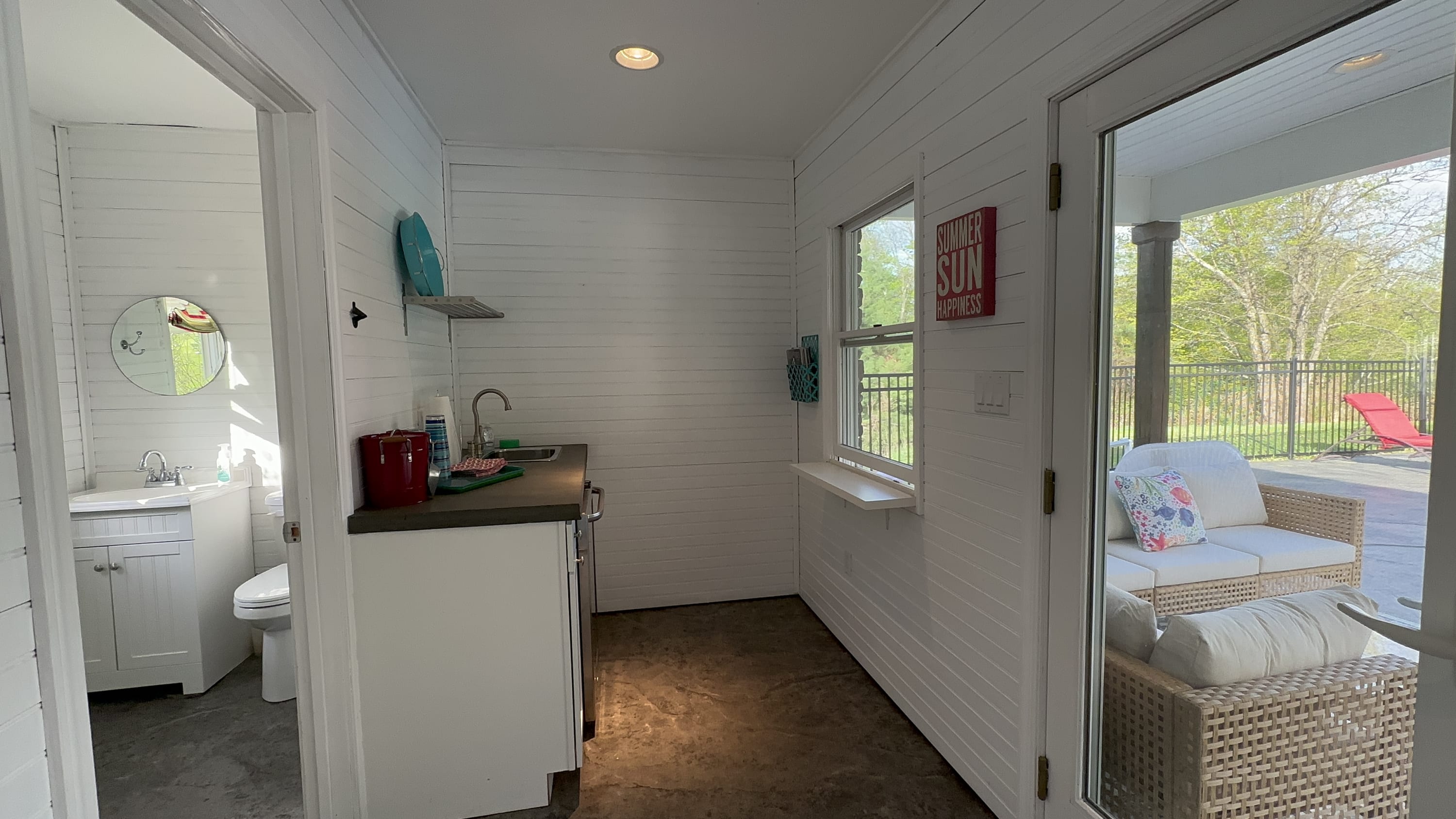 The interior of a pool house features white shiplap walls, a small kitchenette with a sink and dark countertop, and an adjacent bathroom with a white vanity and toilet, with a view to the outdoor patio.
