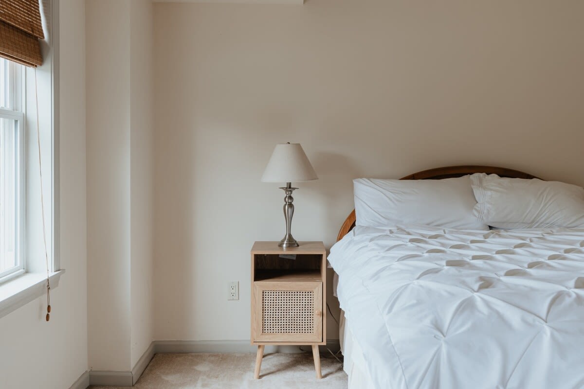 A cozy bedroom featuring a white bed with a tufted duvet, a light wood nightstand with a silver lamp, and a window with a woven blind. The walls are a light neutral color and the floor is carpeted.