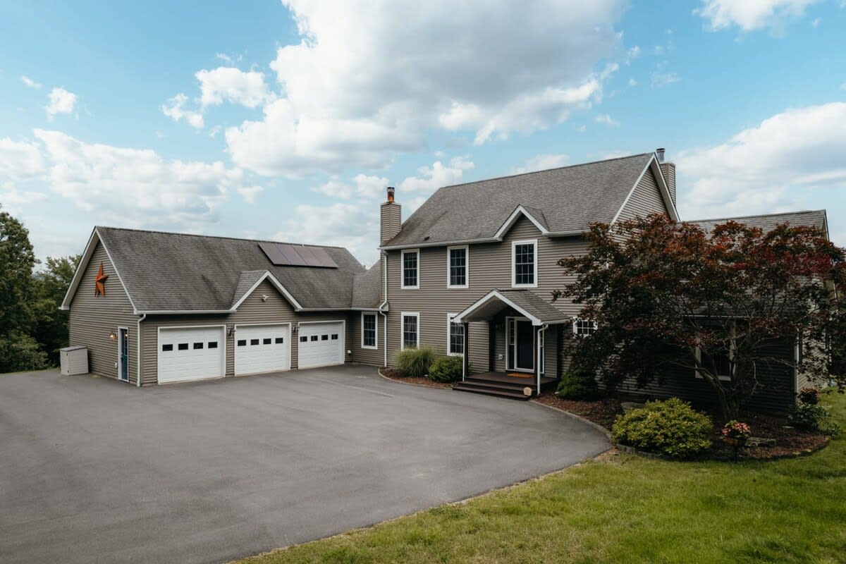 An expansive two-story house with light green horizontal siding and a dark gray shingled roof, featuring a large attached three-car garage with white doors and solar panels. A wide paved driveway curves in front of the property, surrounded by lush green lawn and mature trees under a partly cloudy sky.