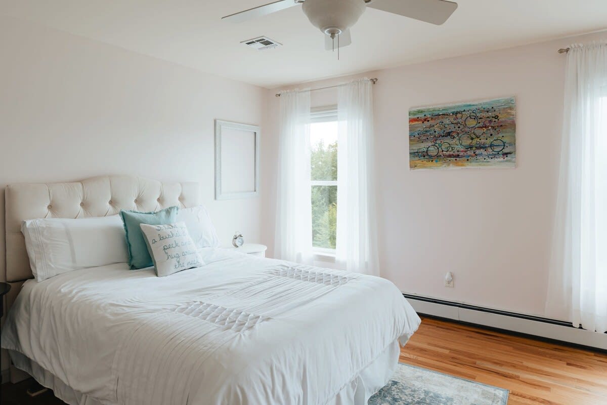 A bright bedroom featuring a white tufted headboard on a queen-sized bed with white bedding, light pink walls, and a window with sheer white curtains, complemented by a ceiling fan.