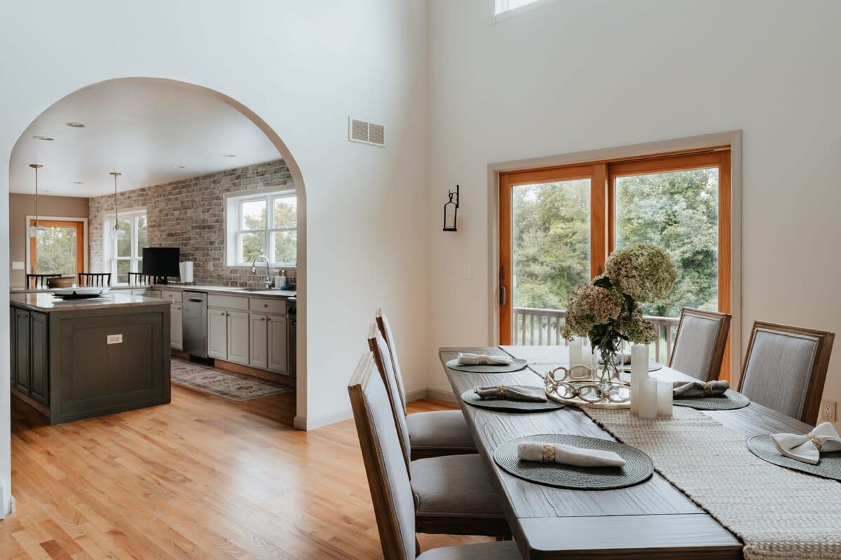 A formal dining area features a long dark wood table set with placemats and a centerpiece, surrounded by upholstered chairs. An arched doorway leads to a modern kitchen with a brick accent wall and an island.
