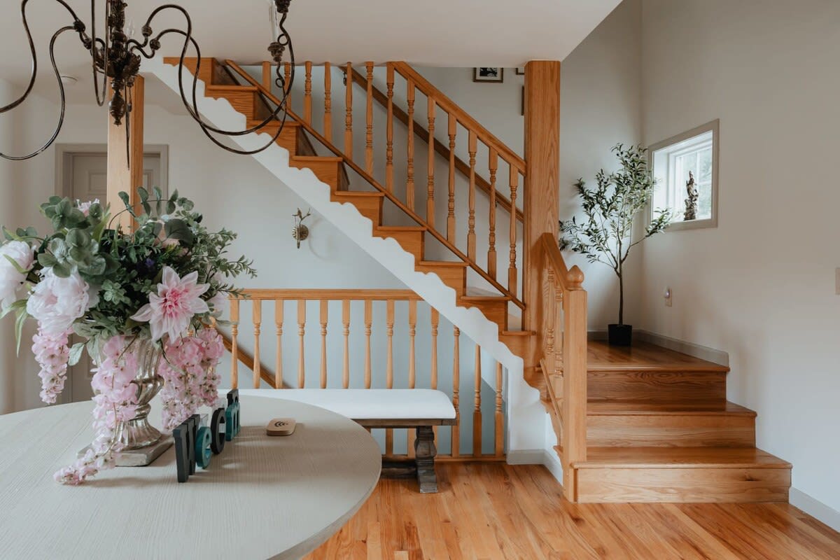 A bright entryway features a wooden staircase with a classic baluster design, leading to upper and lower levels. Hardwood floors extend throughout the space, with a round table and floral arrangement in the foreground.
