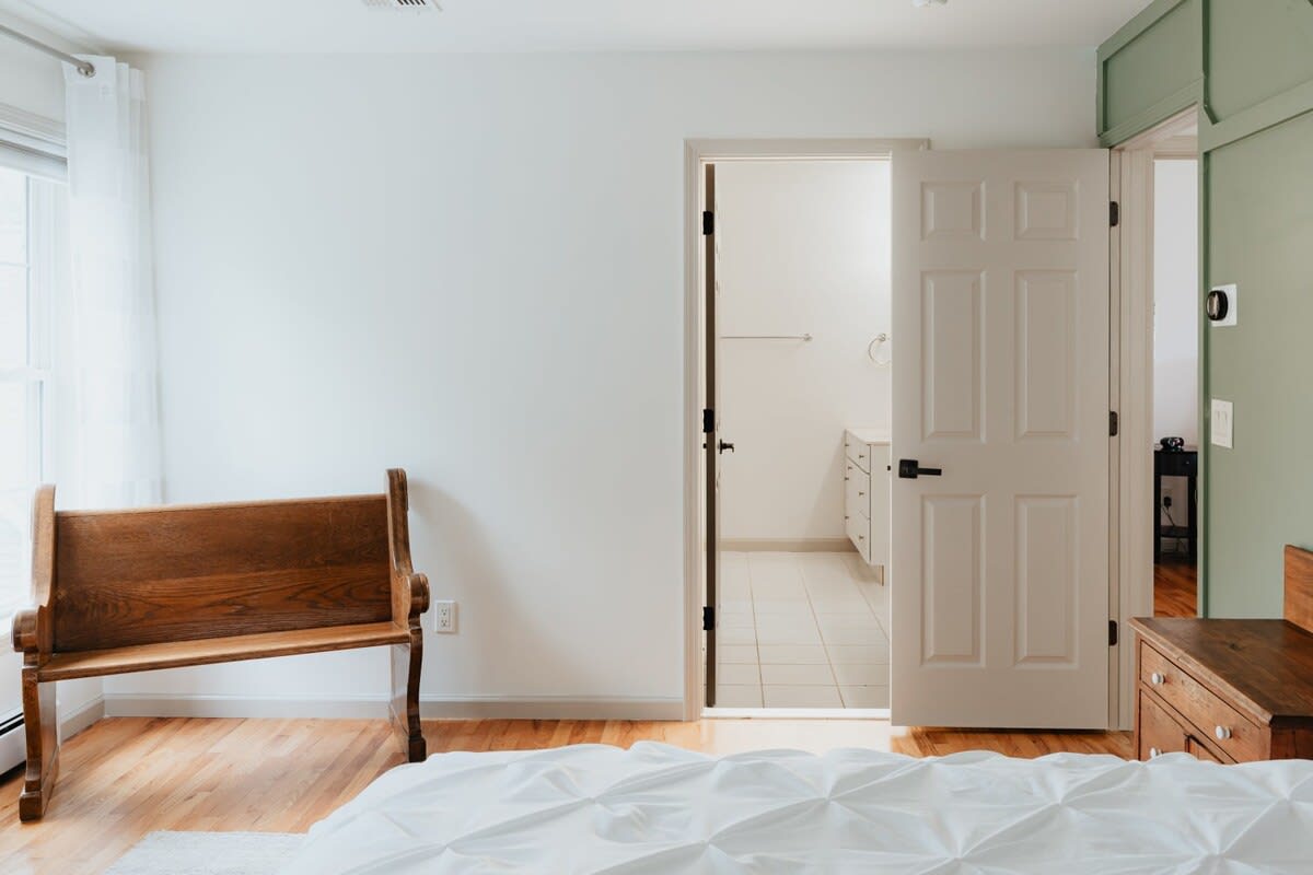 A view of the bedroom showing a rustic wooden bench, an open doorway leading to a tiled bathroom, and a green paneled accent wall, with natural light from a window.