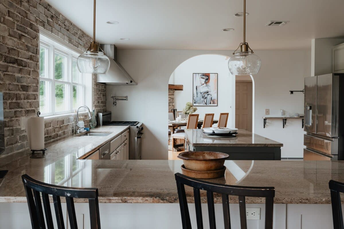 A wide view of the kitchen shows a large island with seating, granite countertops, and a brick backsplash. Pendant lights hang above the island, and stainless steel appliances are visible, with an arched opening to another room.